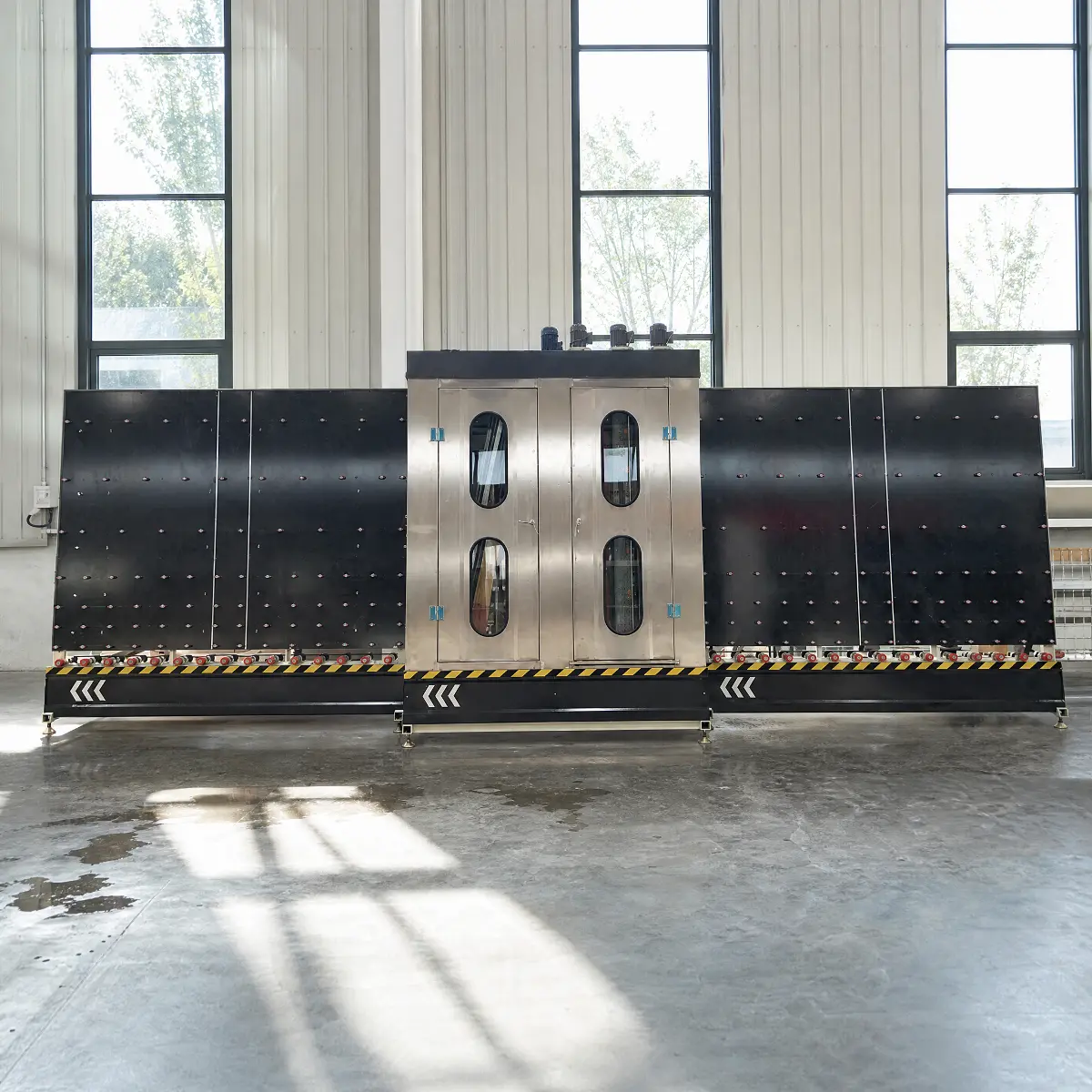 A front-facing view of the vertical glass washing and drying machine. The central section is made of brushed stainless steel with two oval windows, flanked by large black panels with a grid of small studs. The base features a yellow and black hazard-striped trim.